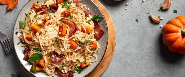 Top View of a Plate of Spaghetti Pasta with Chunks of Roasted Pumpkin & Crispy Bacon. There are Fall Leaves & Decorative Pumpkins on the Tablescape