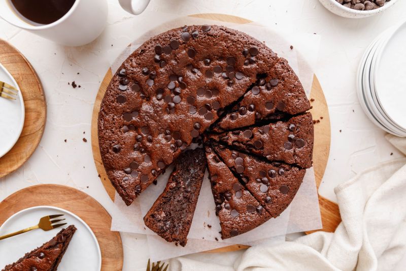 Round Chocolate Espresso Cake Cut into Slices on Round Wooden Cutting Board Surrounded by Plates and Coffee Mugs