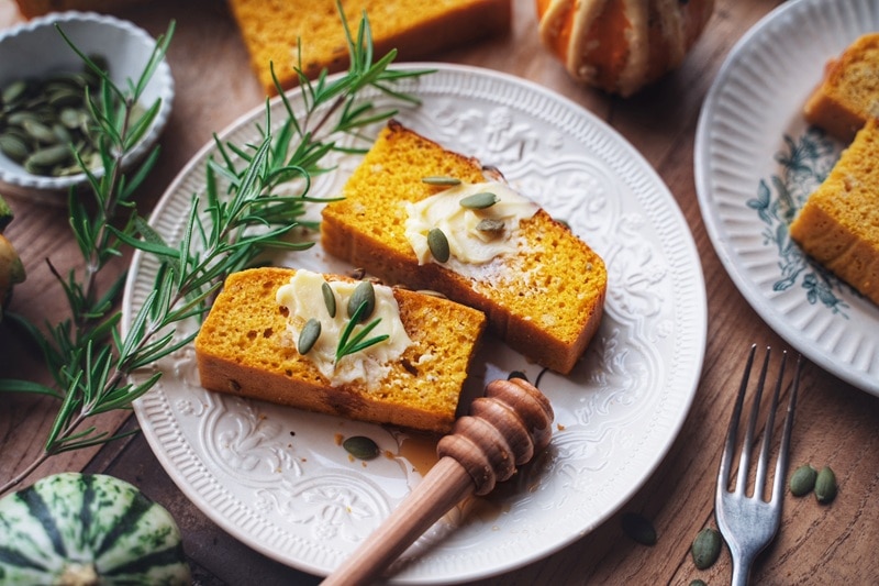 Two Slices of Pumpkin Cornbread Are Topped With Vegan Butter, Pumpkin Seeds and Rosemary.