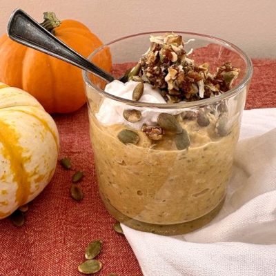 Pumpkin Pie Overnight Oats in a Glass Jar, Topped with a Crumble of Chopped Dates, Pumpkin Seeds and Coconut, along with a Dollop of Yogurt. Two Autumnal Gourds Sit on the Table Beside it