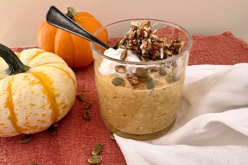 Pumpkin Pie Overnight Oats in a Glass Jar, Topped with a Crumble of Chopped Dates, Pumpkin Seeds and Coconut, along with a Dollop of Yogurt. Two Autumnal Gourds Sit on the Table Beside it