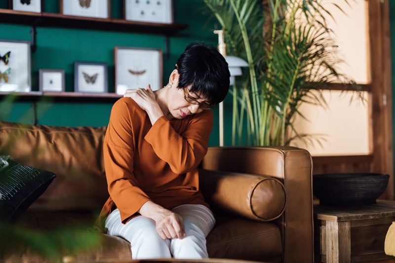 A Woman Sitting on a Couch Massages Her Shoulder, Representing the Question, "What Are Muscle Knots?"