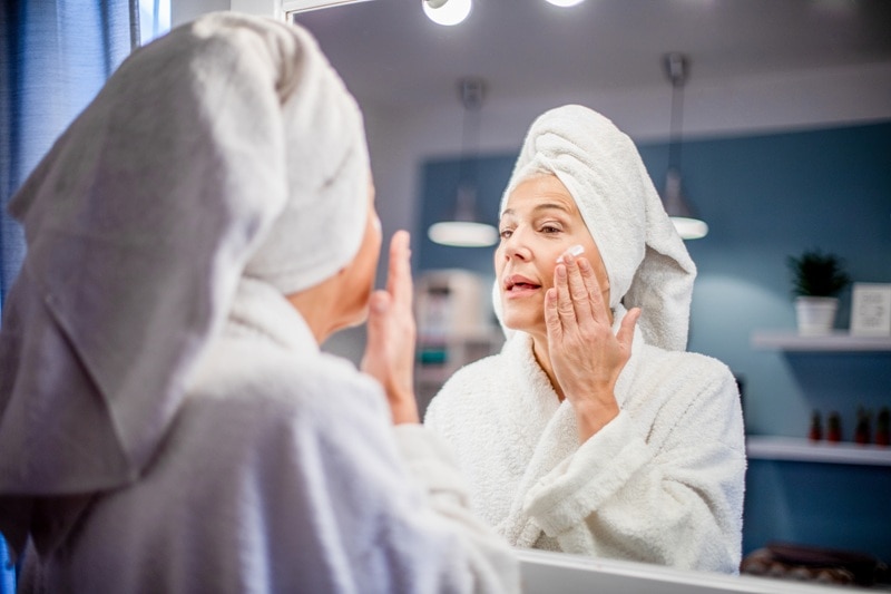 A Woman in a Robe Applies Cream to Her Cheek, Representing What to Do After Microneedling.