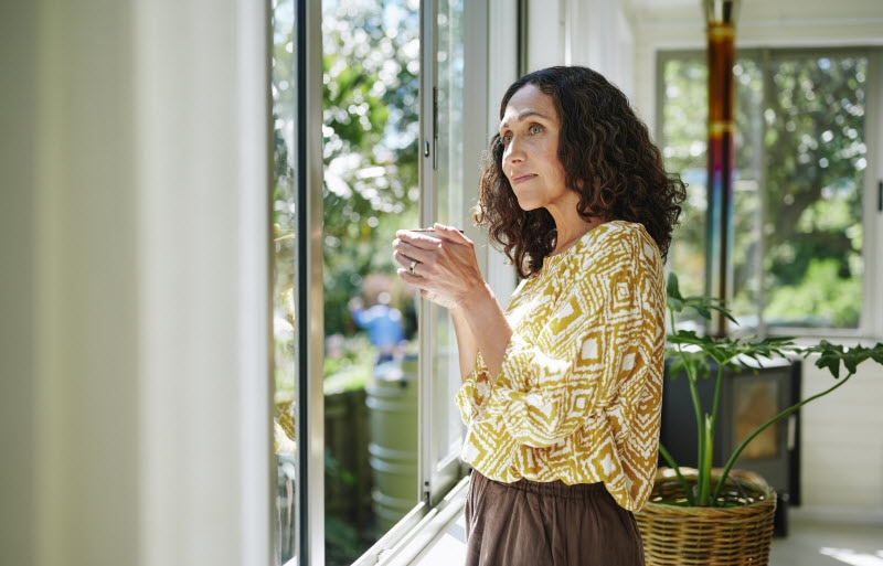Pensive-Looking 50-Year-Old Woman Looking Out Living Room Window Wondering About the Connection Between Breast Cancer and Menopause