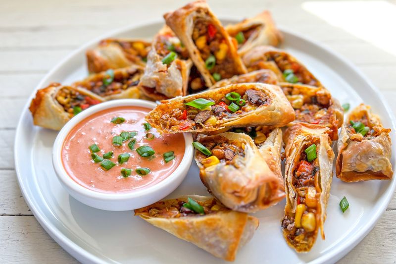 Diagonally Sliced Vegan Southwestern Egg Rolls Filled with Corn, Black Beans, Lentils, Diced Red Peppers & Scallions Surround a Bowl of Salsa-Ranch Dipping Sauce
