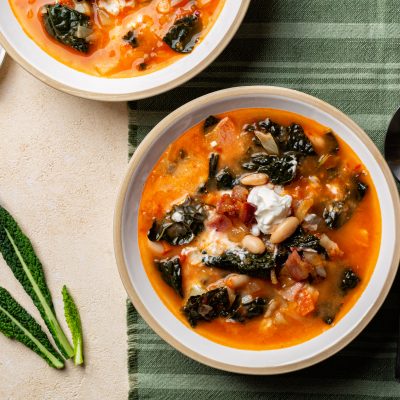 Two Bowls of Tuscan Soup With Kale, White Beans, and Tomato Broth on a Green Striped Cloth With Spoons and Fresh Kale Leaves Beside Them.