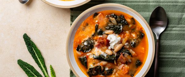 Two Bowls of Tuscan Soup With Kale, White Beans, and Tomato Broth on a Green Striped Cloth With Spoons and Fresh Kale Leaves Beside Them.