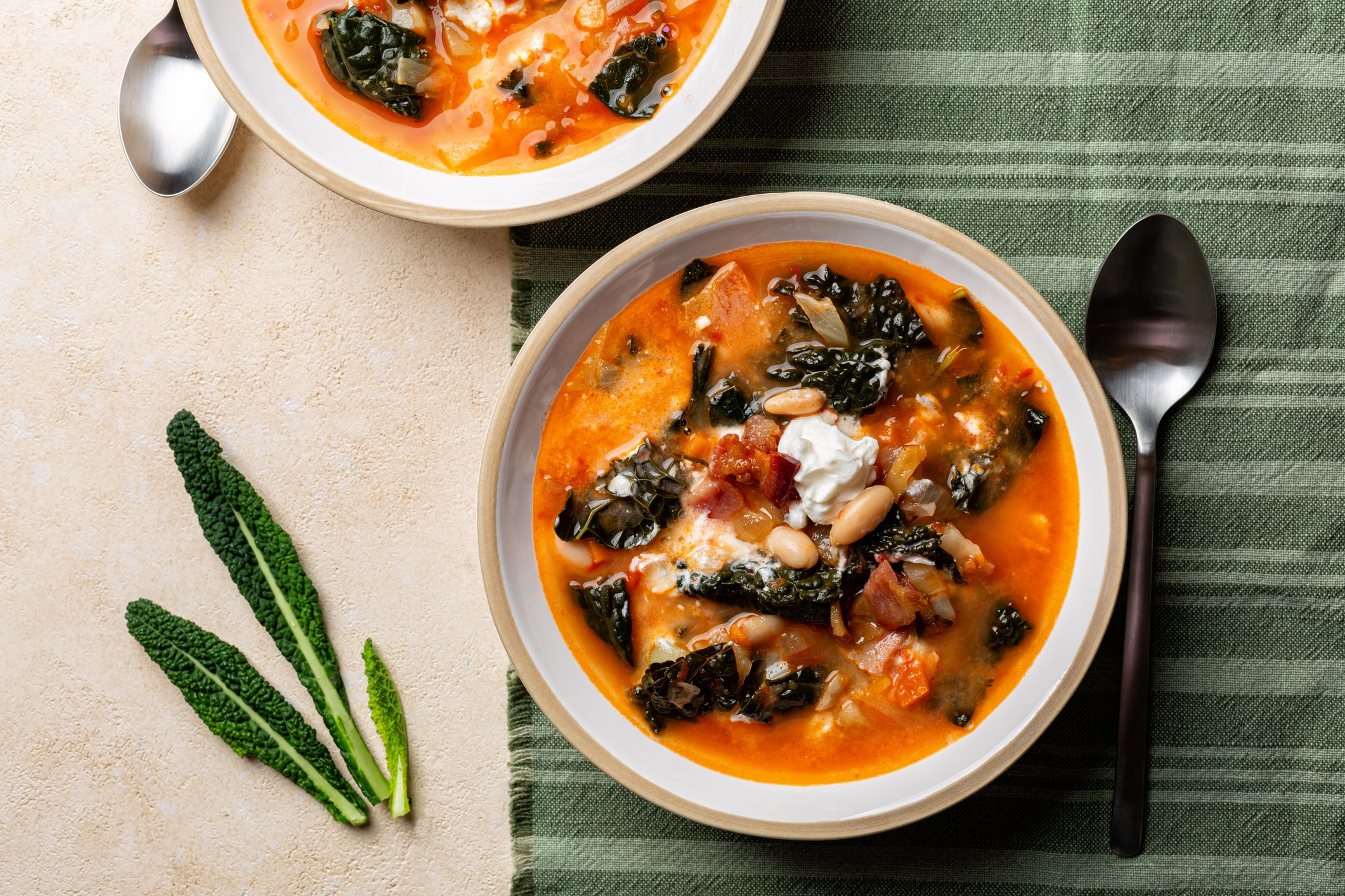 Two Bowls of Tuscan Soup With Kale, White Beans, and Tomato Broth on a Green Striped Cloth With Spoons and Fresh Kale Leaves Beside Them.