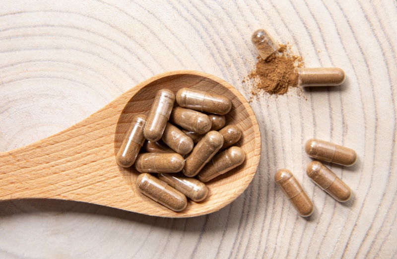 Concept of How to Make Your Own Supplements Represented by Capsules Filled with Beige Powder on Wooden Spoon on Wood-Grain Background