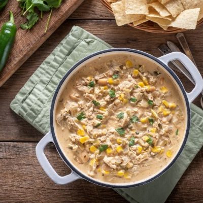 Instant Pot White Chicken Chili in Handled Enamelware Bowl on a Wooden Plank Tabletop With a Linen Napkin, Tortilla Chips and Lime Wedges Nearby