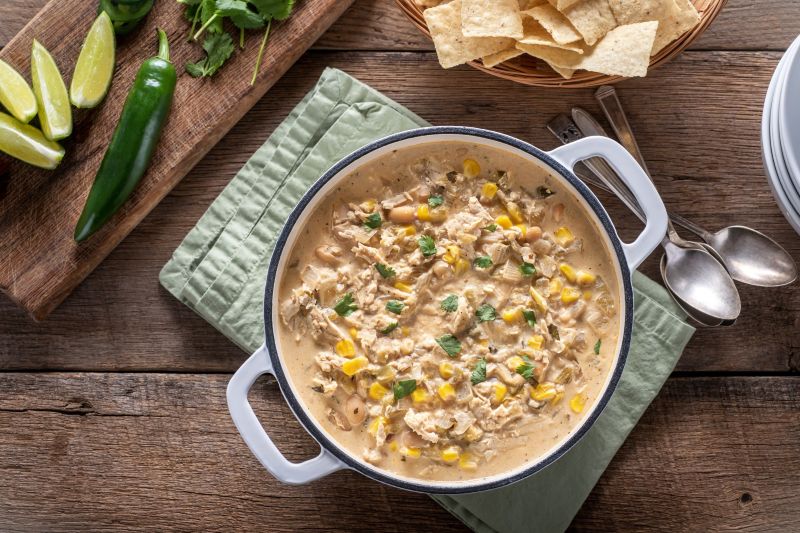 Instant Pot White Chicken Chili in Handled Enamelware Bowl on a Wooden Plank Tabletop With a Linen Napkin, Tortilla Chips and Lime Wedges Nearby