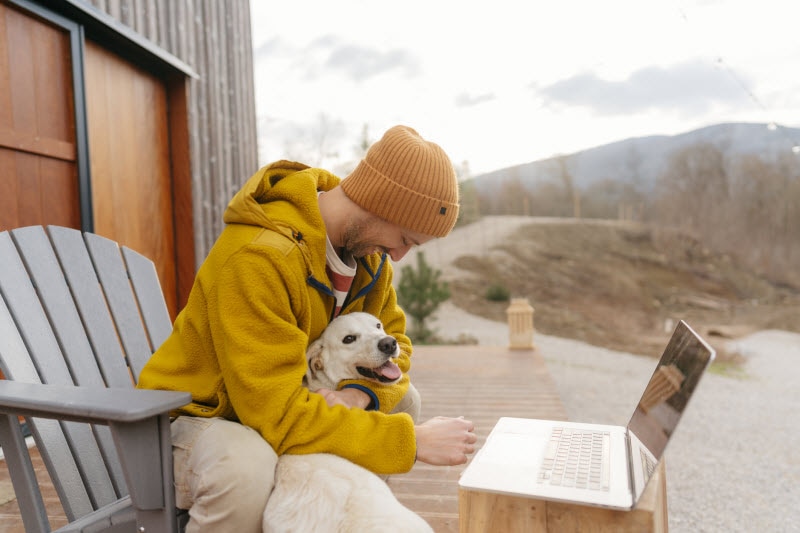 Concept of Is Cold Weather Bad for Your Lungs Represented by Guy in Orange Hat Holding Dog While Working on Laptop Outdoors in Autumn