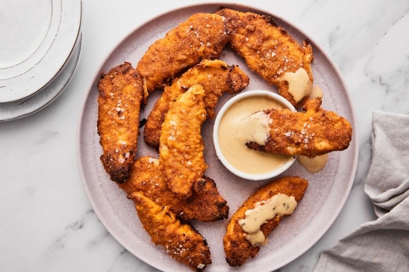 Plate of Maple-Dijon Chicken Tenders Served with Creamy Maple Mustard Dipping Sauce on a Light Pink Plate with a Napkin and Stack of Plates Nearby