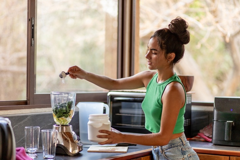 A Young Woman Adds a Scoop of Protein Powder to a Blender With Fruits and Vegetables, Representing a Protein Powder Without Artificial Sweetener.