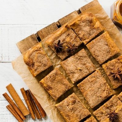 Top View of Pumpkin Blondies Cut Into Squares on Parchment Paper with Cinnamon Sticks, Star Anise, and Jar of Caramel Sauce on White Wooden Table