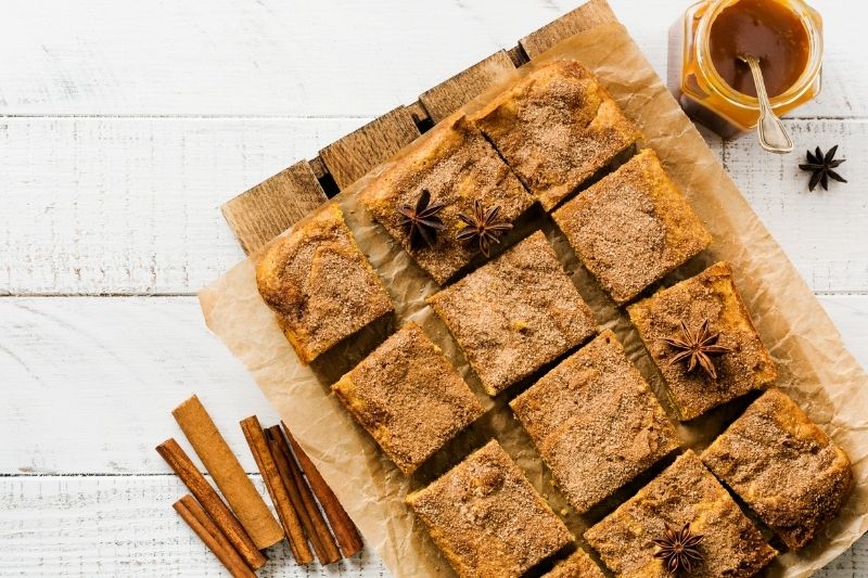 Top View of Pumpkin Blondies Cut Into Squares on Parchment Paper with Cinnamon Sticks, Star Anise, and Jar of Caramel Sauce on White Wooden Table