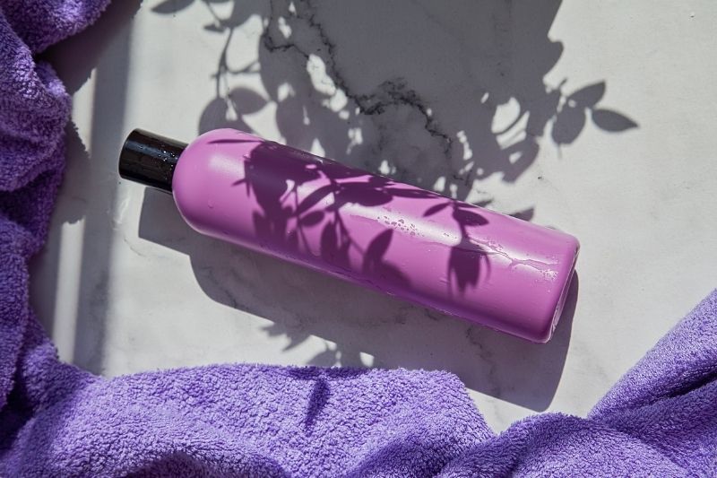 Purple Shampoo Bottle on White Marble Surface with Purple Towel and Leaf Shadows