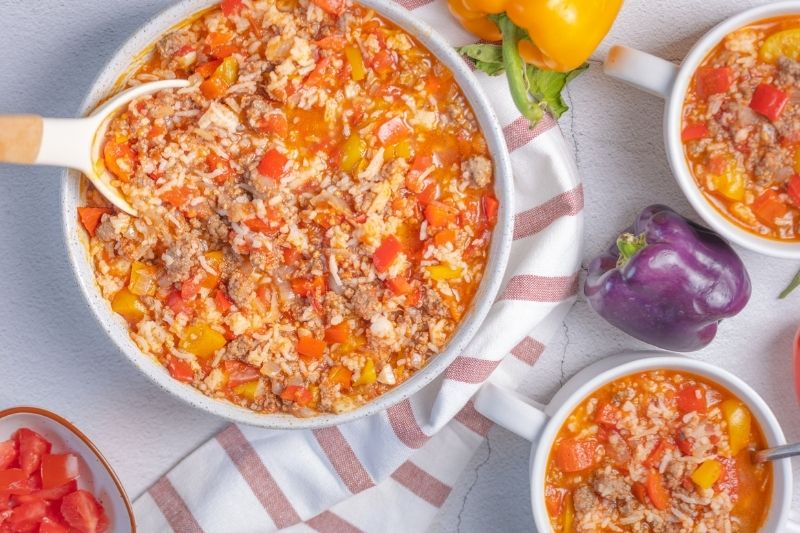 Bowl And Mugs Of Hearty Stuffed Pepper Soup Made With Ground Beef, Rice, Diced Tomatoes, And Colorful Bell Peppers, Served On A Striped Towel With Fresh Peppers
