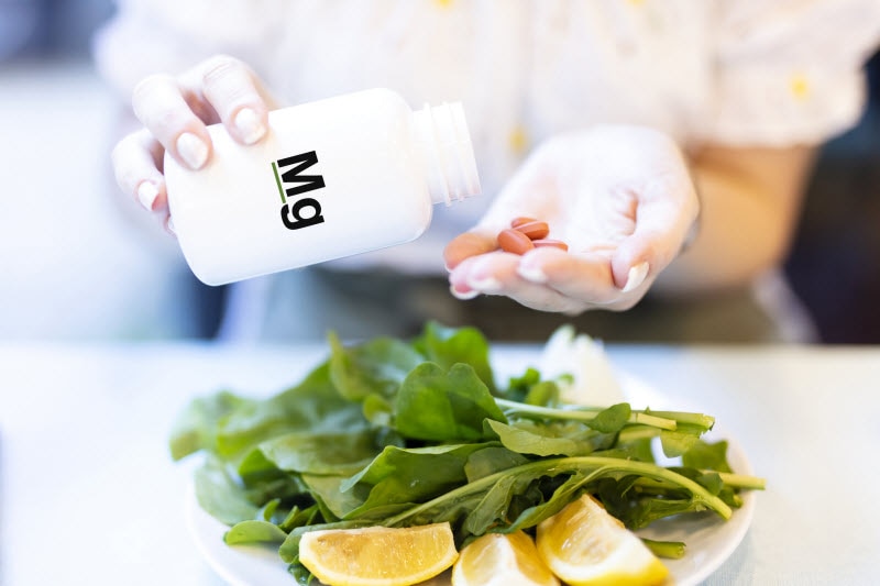 Concept of Types of Magnesium Represented by Woman Pouring Capsules into Hand from White Bottle Over Plate of Spinach Salad