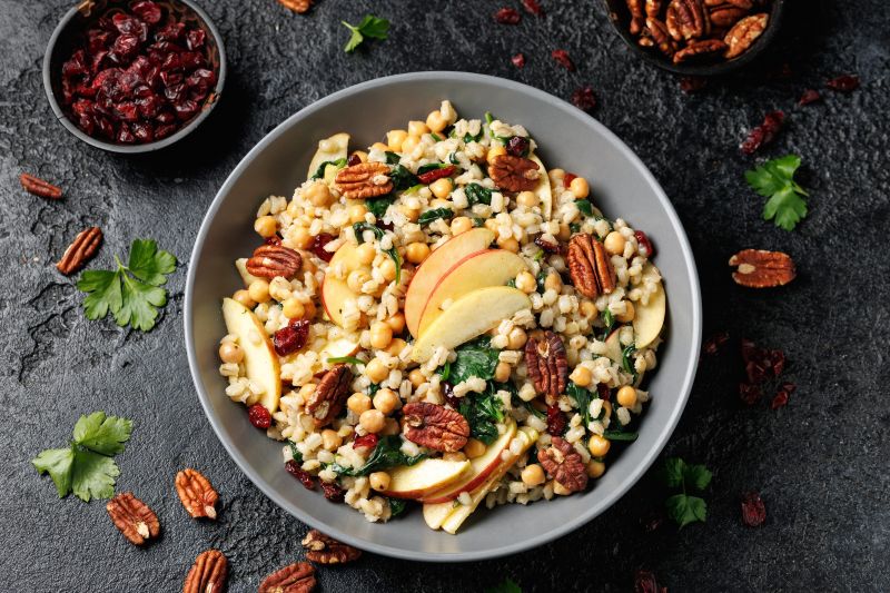 Pearl Barley, Kale & Apple Salad with Pecan Halves in Big Bowl on Slate Countertop Dotted with Cranberries and Pecan Halves