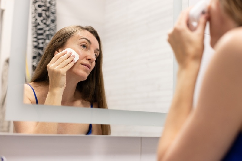 A Woman Applies Waterless Skincare to Her Face in the Mirror, Representing the Question, "What is Waterless Skincare?"