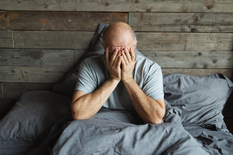 Man With Insomnia Sitting In Bed Rubbing Eyes Wondering Can Lack of Sleep Cause Dementia