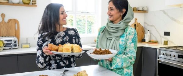 Healthy Fall Traditions Around the World Two Women Sharing Homemade Dishes in a Bright Modern Kitchen, Smiling at Each Other While Holding Plates of Traditional Food on a Counter Filled With Naan, Curries, and Pastries