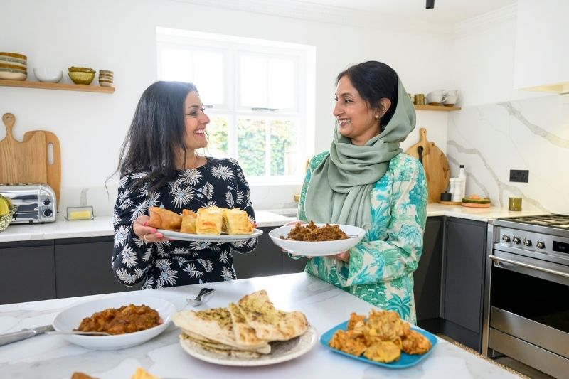 Two Women Sharing Homemade Dishes in a Bright Modern Kitchen, Smiling at Each Other While Holding Plates of Traditional Food on a Counter Filled With Naan, Curries, and Pastries