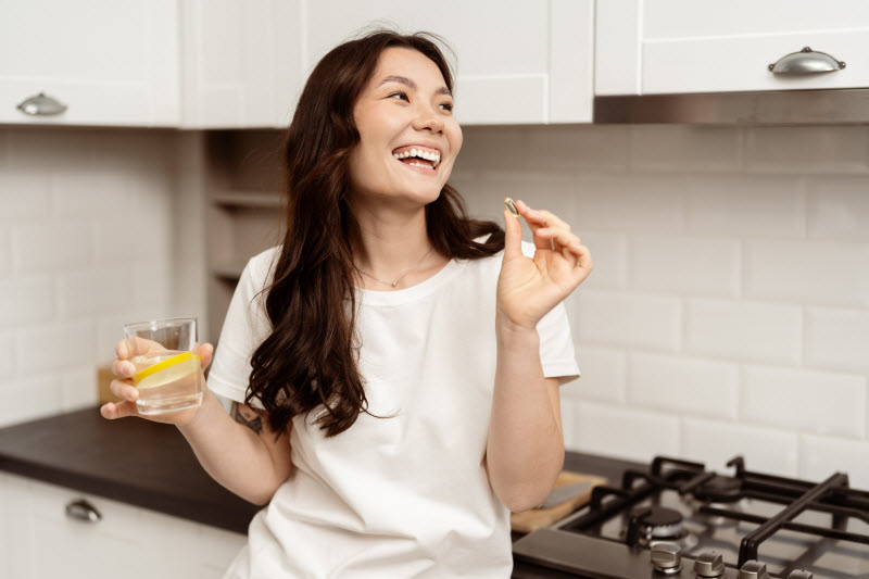 Concept of How Do Digestive Enzymes Work Represented by Smiling Woman in Kitchen Taking Supplement With Glass of Water