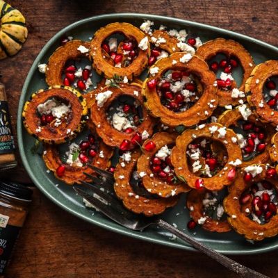 Circles of Roasted Delicata Squash Sprinkled With Feta Cheese and Pomegranate Seeds on an Oval Serving Platter Next to Bottles of Frontier Co-op Spices