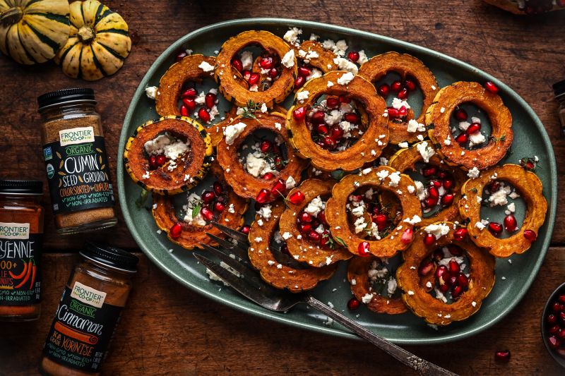 Circles of Roasted Delicata Squash Sprinkled With Feta Cheese and Pomegranate Seeds on an Oval Serving Platter Next to Bottles of Frontier Co-op Spices