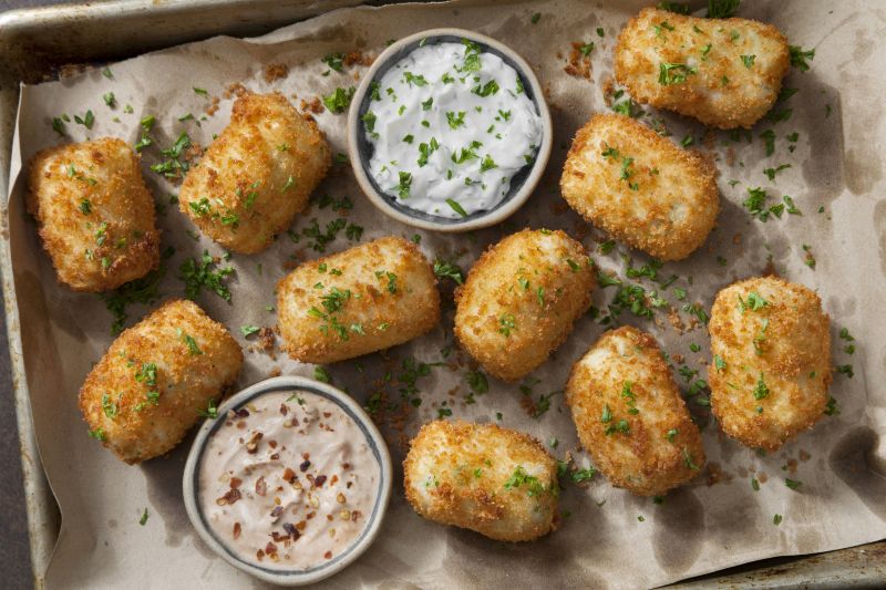 Breaded Mashed Potato Croquettes on Parchment-Lined Sheet Pan with Creamy Dipping Sauces in Two Bowls