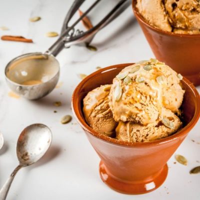 Vegan Pecan Pumpkin Pie Ice Cream in Serving Dish With Pumpkin Seeds Sprinkled on Top, Spoons and an Ice Cream Scoop Adorn the Counter