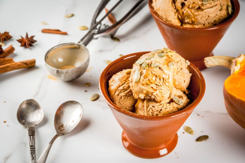 Vegan Pecan Pumpkin Pie Ice Cream in Serving Dish With Pumpkin Seeds Sprinkled on Top, Spoons and an Ice Cream Scoop Adorn the Counter