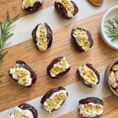 Vegan Stuffed Medjool Dates With Rosemary Tofu Ricotta, Hot Agave “Honey,” and Chopped Pistachios Arranged on a Wooden Serving Board With Fresh Rosemary and Pistachios