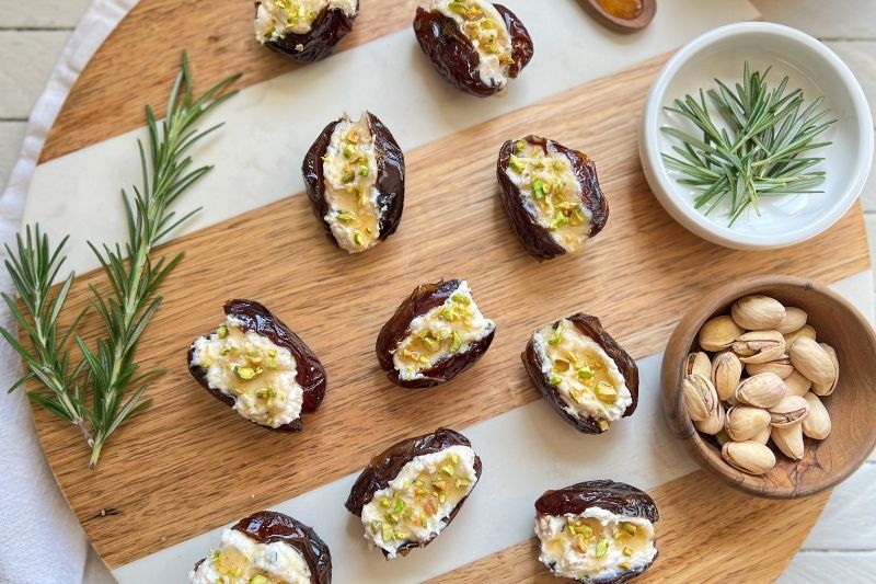 Vegan Stuffed Medjool Dates With Rosemary Tofu Ricotta, Hot Agave “Honey,” and Chopped Pistachios Arranged on a Wooden Serving Board With Fresh Rosemary and Pistachios