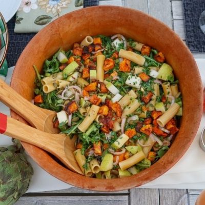 Colorful Winter Pasta Salad With Roasted Butternut Squash, Arugula, Diced Pear, Red Onion, and Toasted Pumpkin Seeds in a Wooden Serving Bowl With Red-Tipped Wooden Tongs, Surrounded by Fresh Apples, Pears, and Grapes on an Outdoor Table Setting