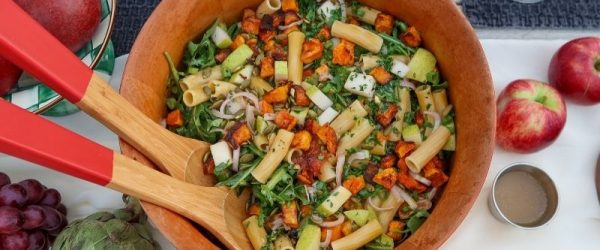 Colorful Winter Pasta Salad With Roasted Butternut Squash, Arugula, Diced Pear, Red Onion, and Toasted Pumpkin Seeds in a Wooden Serving Bowl With Red-Tipped Wooden Tongs, Surrounded by Fresh Apples, Pears, and Grapes on an Outdoor Table Setting