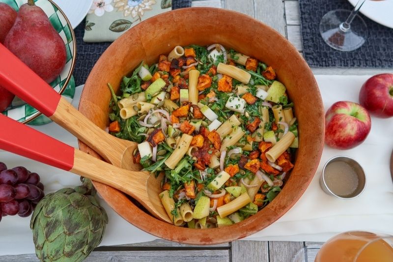 Colorful Winter Pasta Salad With Roasted Butternut Squash, Arugula, Diced Pear, Red Onion, and Toasted Pumpkin Seeds in a Wooden Serving Bowl With Red-Tipped Wooden Tongs, Surrounded by Fresh Apples, Pears, and Grapes on an Outdoor Table Setting
