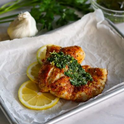 Seasoned Baked Cod Drizzled With Gremolata Sauce on a Sheet Pan Surrounded by Lemon Slices and a Bulb of Fresh Garlic