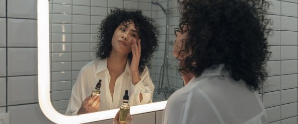 Benefits of Vitamin C Serum A Woman Applies a Serum While Looking in the Bathroom Mirror, Representing the Benefits of Vitamin C Serum.