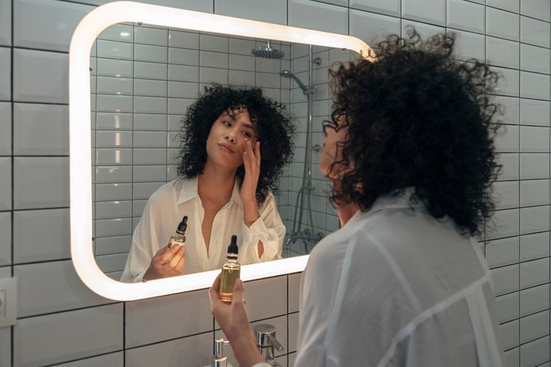 A Woman Applies a Serum While Looking in the Bathroom Mirror, Representing the Benefits of Vitamin C Serum.