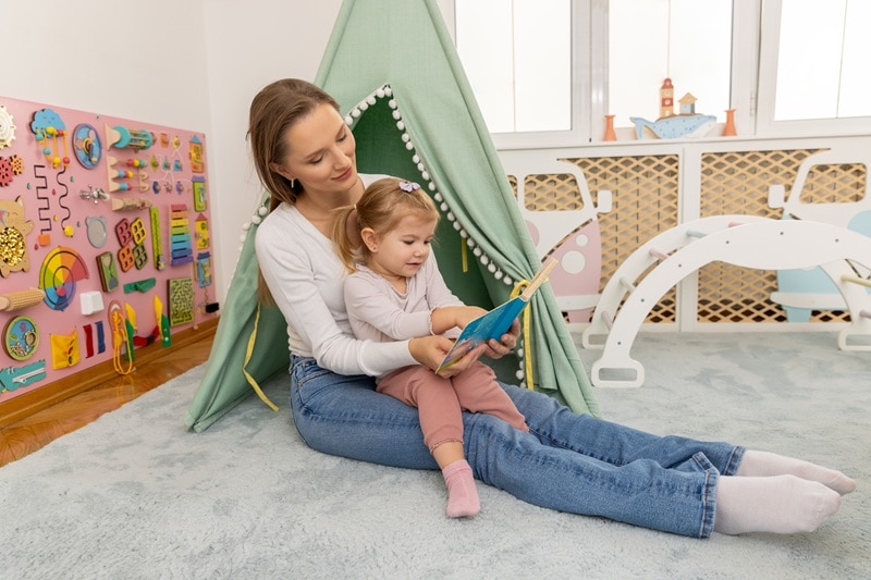 A Mother Sits With Her Toddler and Reads Near a Small Tent and Sensory Games, Representing Calming Corner Ideas.