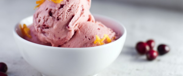 A Small Bowl of Cranberry Nice Cream with Curls of Orange Zest Sprinkled on it, Whole Fresh Cranberries are Strewn About on the Counter