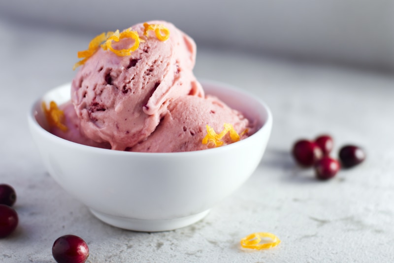A Small Bowl of Cranberry Nice Cream with Curls of Orange Zest Sprinkled on it, Whole Fresh Cranberries are Strewn About on the Counter