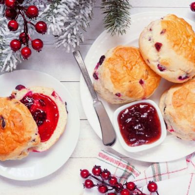Fresh cranberry scones with jam on round white plates. Top view table scene on a white wood background. Christmas fur tree boughs and red berries are scattered about.