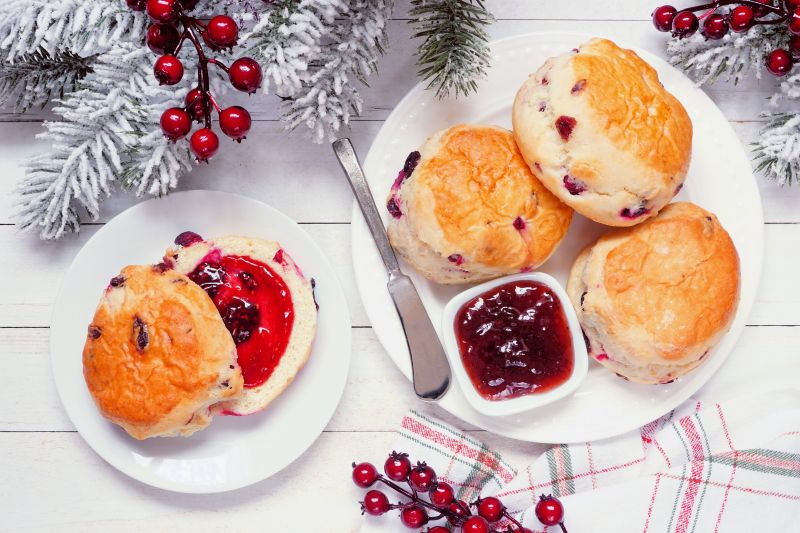 Fresh cranberry scones with jam on round white plates. Top view table scene on a white wood background. Christmas fur tree boughs and red berries are scattered about.