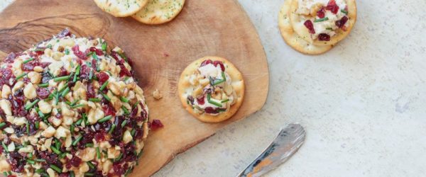 Cranberry-Walnut Cheeseball_Resized Image_Liana Werner-Gray A Round Vegan Holiday Cheese Ball Covered in Cranberries and Walnuts on Wood Charcuterie Board With a Cheese Spreader Knife and Crackers Nearby