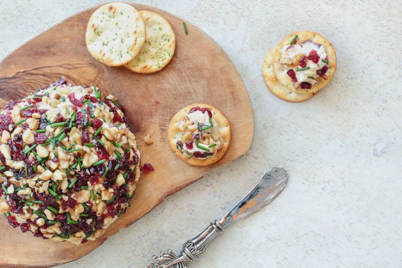 A Round Vegan Holiday Cheese Ball Covered in Cranberries and Walnuts on Wood Charcuterie Board With a Cheese Spreader Knife and Crackers Nearby