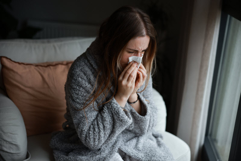 Flu Prevention 101 - How to Lower Your Risk of Infection and Complications Woman Blowing Nose on Couch After Contracting Illness Because She Didn't Follow Flu Prevention Tips